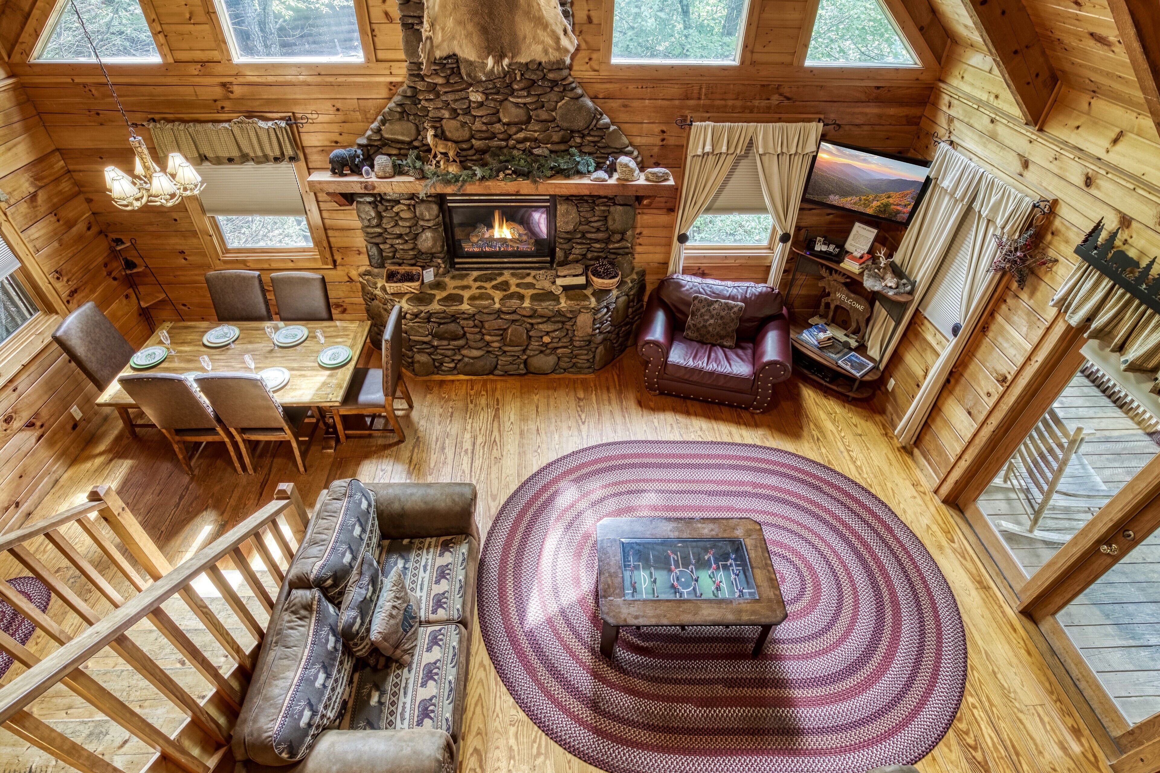 A cozy cabin interior with a stone fireplace, wooden furniture, and a large window overlooking a forest.