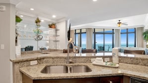 A modern kitchen with granite countertops, a stainless steel sink, and a built-in dishwasher. There is a seating area with a view of the ocean.