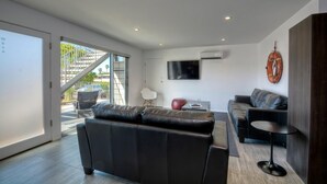 A modern living room with a large sliding glass door leading to a balcony, a flat-screen TV mounted on the wall, and a dark leather sofa. - Wave (2 bedroom / 2 bath) (Carlsbad)
