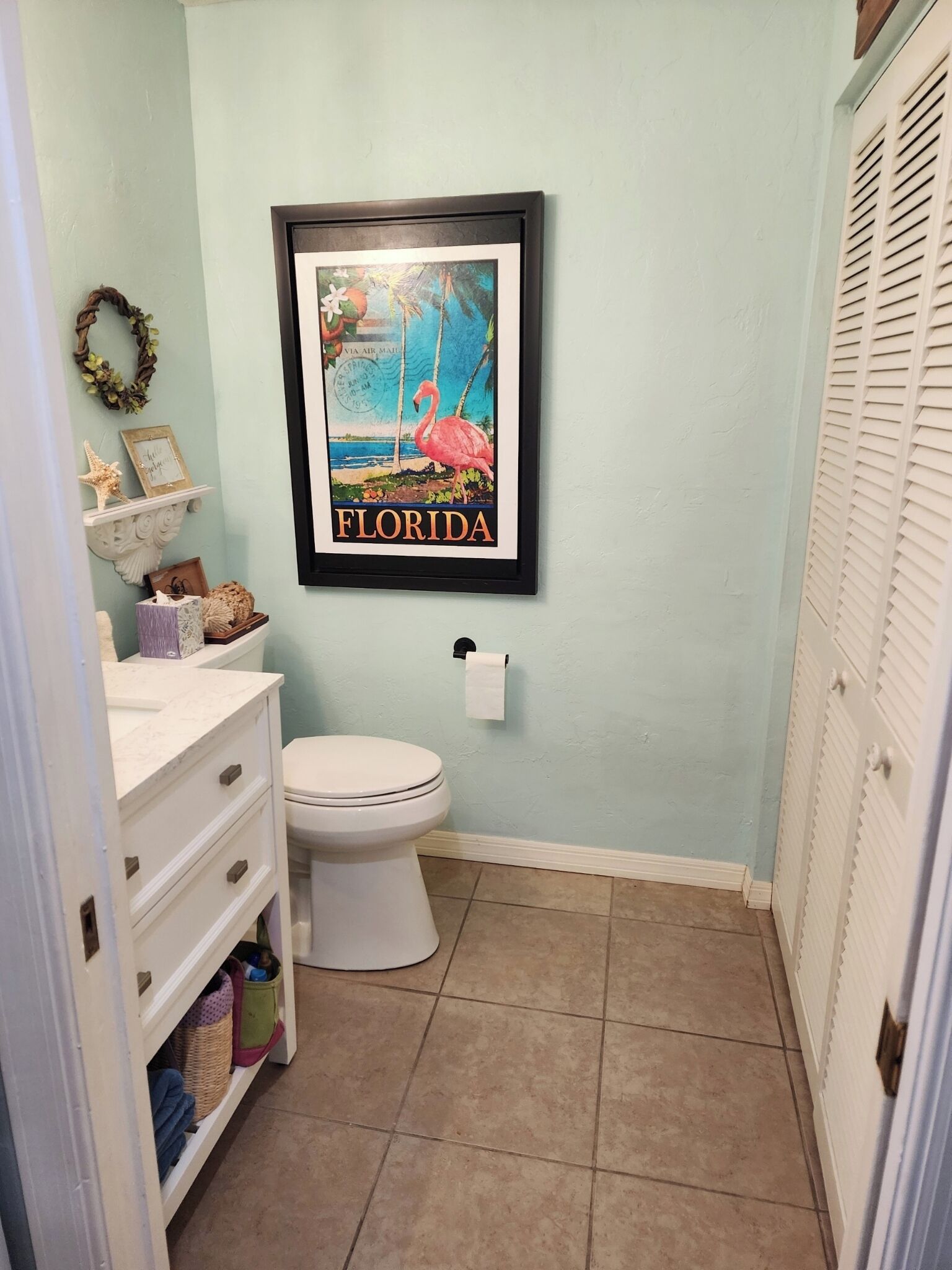 Un baño con inodoro, lavabo con tocador, un cuadro enmarcado de una playa de Florida y un perchero.