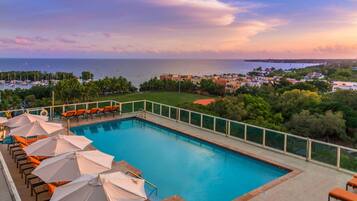Ein Swimmingpool mit Liegestühlen und Sonnenschirmen, umzäunt, mit Blick auf das Meer und Gebäude im Hintergrund.