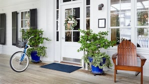 Un porche avec un vélo bleu, deux plantes en pot, une chaise en bois et une porte sur laquelle est affiché le mot « BONJOUR ».