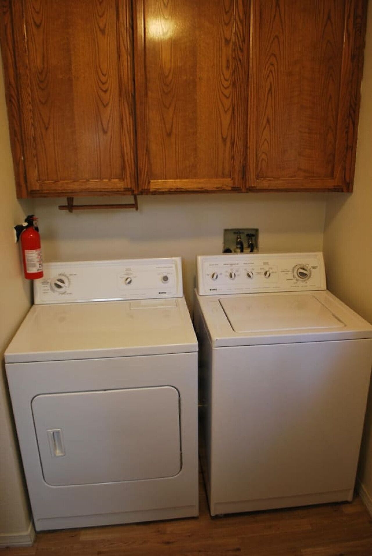 A laundry area with two white front-loading washers and dryers, wooden cabinets above, and a fire extinguisher on the left.