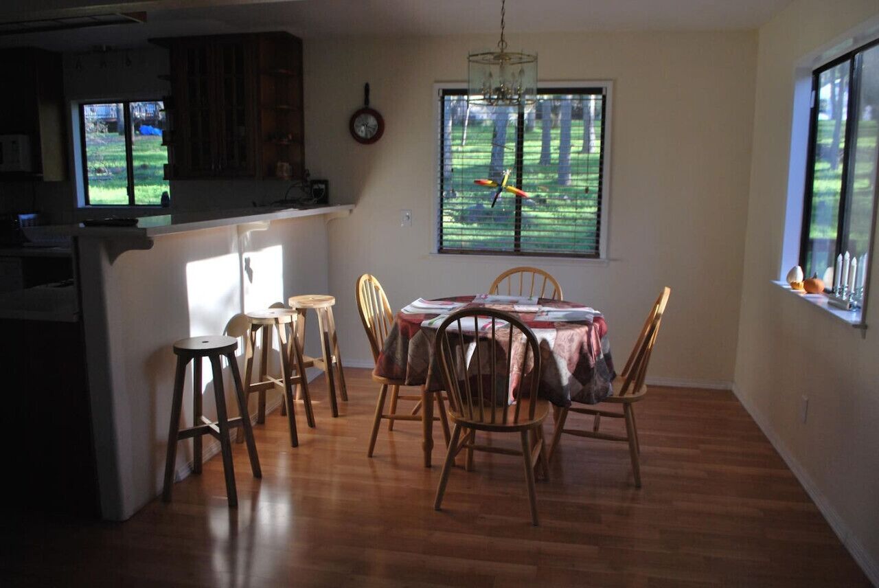A dining area with a table set for four, wooden chairs, a window with a view of trees, and a kitchen area with wooden cabinets and a clock on the wall.