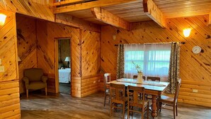 Une cabane en bois chaleureuse, avec un coin repas comprenant une table et des chaises, un petit fauteuil et un lit en arrière-plan.