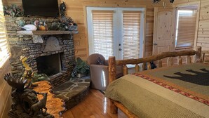 A log cabin interior with a stone fireplace, wooden furniture, and a bed with a patterned bedspread.