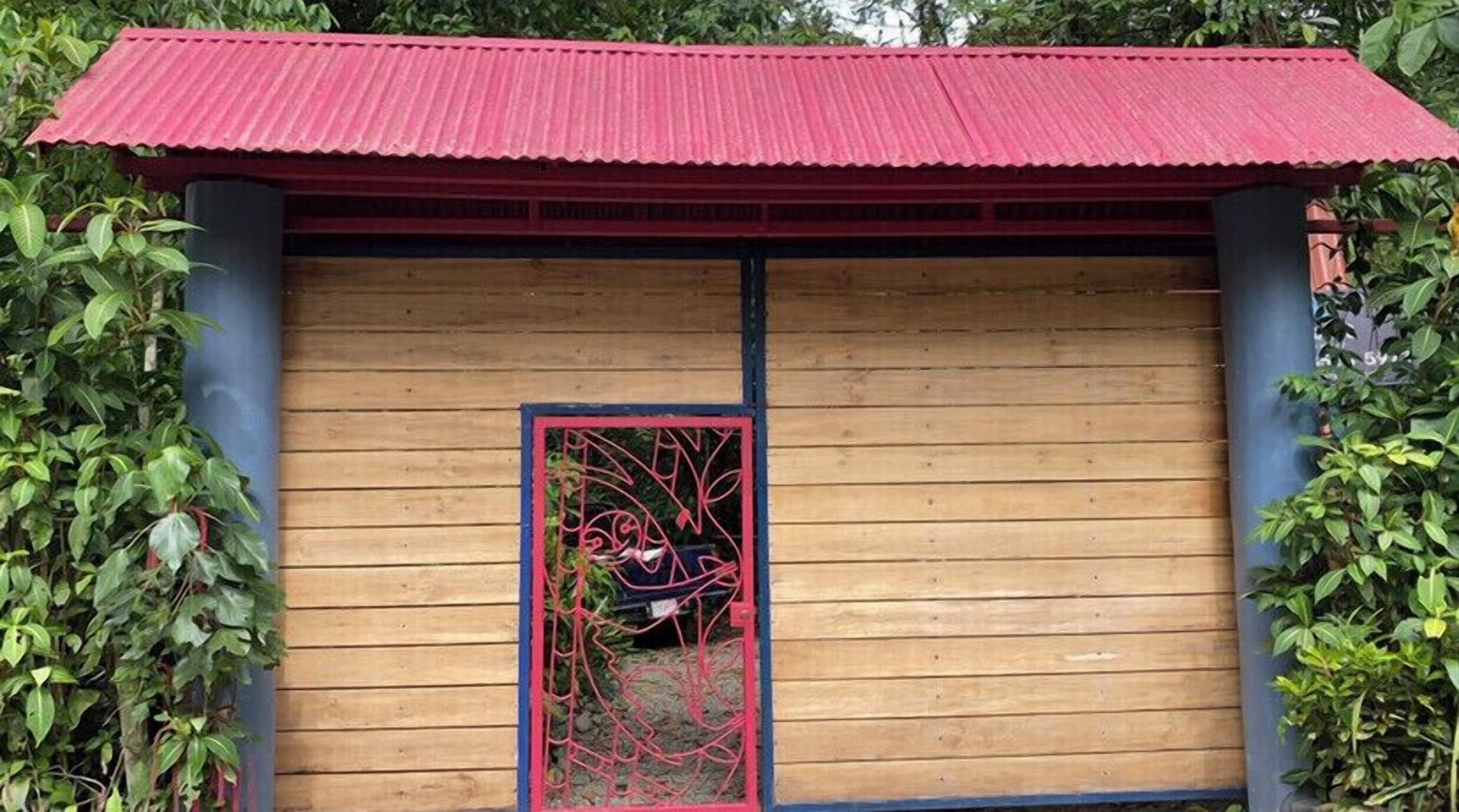 Una estructura de madera con techo de láminas rojas y puerta azul con reja metálica decorativa.