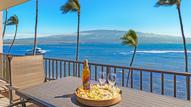 Eine Holzterrasse mit Tisch, einer Flasche und zwei Gläsern, mit Blick auf einen Strand mit Palmen und einem Boot in der Ferne.