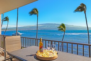 A wooden deck with a table, a bottle, and two glasses, overlooking a beach with palm trees and a boat in the distance. - Two Bedroom Oasis!  (Maalaea)