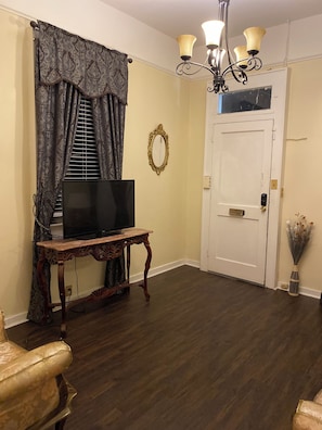 Uma sala de estar com piso de madeira escura, uma televisão sobre uma mesa de madeira, uma cortina estampada, um espelho decorativo e um lustre.