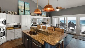 A spacious kitchen with granite countertops, white cabinetry, and a large island with a sink. There are pendant lights, a ceiling fan, and artwork on the walls.