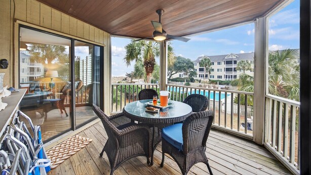 An outdoor deck with wicker furniture, a glass table, and a view of a pool and buildings. - Beautiful Modern Villa with OCEAN VIEWS! Screened Porch, Pool Access! (Isle of Palms)