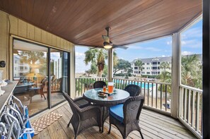 An outdoor deck with wicker furniture, a glass table, and a view of a pool and buildings.
