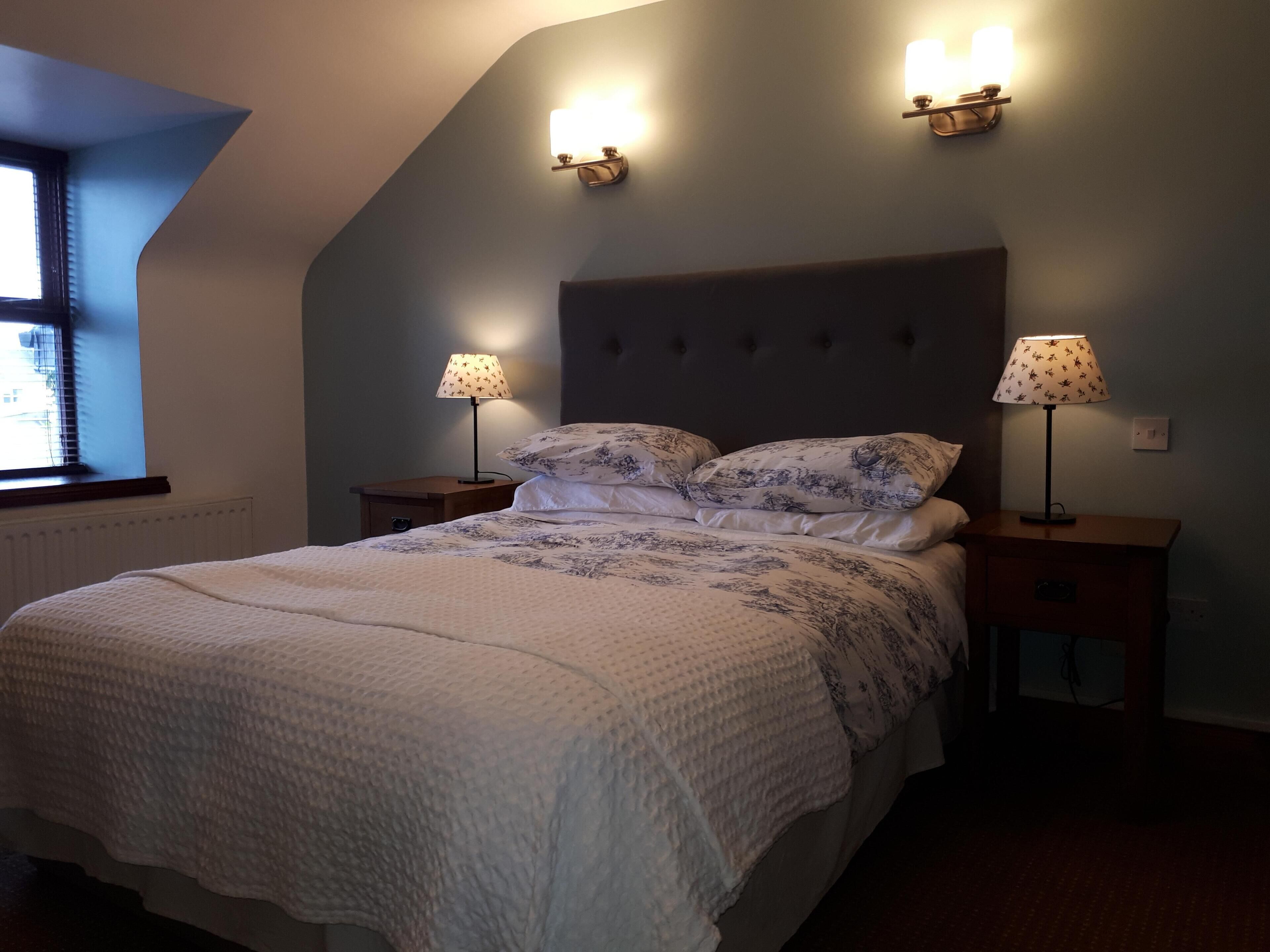 A bedroom with a large bed, two bedside lamps, and a window with blinds.