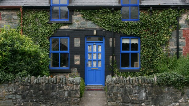 Un bâtiment en pierre avec une porte noire et des fenêtres à cadre bleu, entouré de verdure.