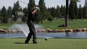 Ein Golfplatz mit einem Wasserhindernis, Bäumen und einem Gebäude im Hintergrund.