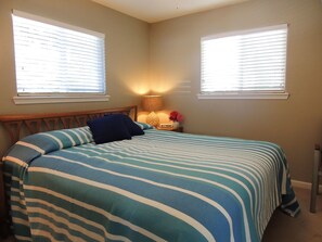 A bedroom with a bed, two windows, a nightstand with a lamp, and a vase with flowers.