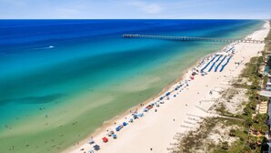 An aerial view of a beach with a pier extending into the ocean, rows of beach umbrellas, and a sandy shoreline.