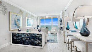 A modern living room with a large painting of water, a white sofa, a dining area with bar stools, and a view of the ocean through large windows.