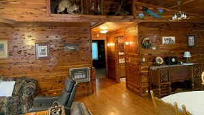 A cozy cabin interior with wooden walls, a fireplace, a dining table, and a loft area with a raccoon. - Beautiful Log Cabin. Sleeps 2-8. Reds Landing boat ramp .5 mi away. A+ fishing. (Norfork)