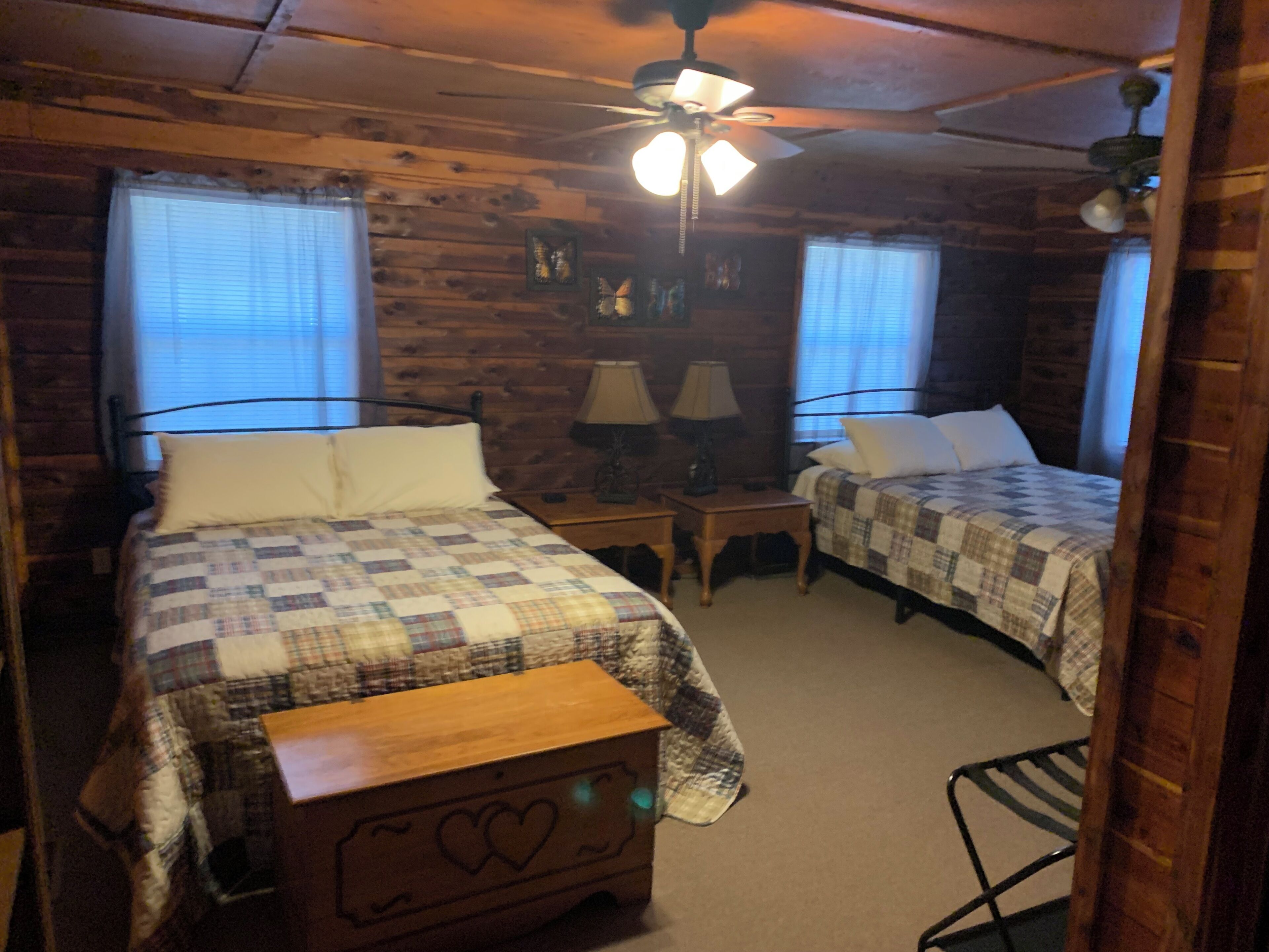 A cabin-style room with two beds, a wooden bench, a ceiling fan, and a small table with lamps.