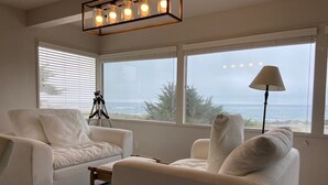 Ein Wohnzimmer mit zwei weißen Sofas, einem Couchtisch, einem Stativ und einer Lampe. Große Fenster bieten Blick auf das Meer und Gebäude.