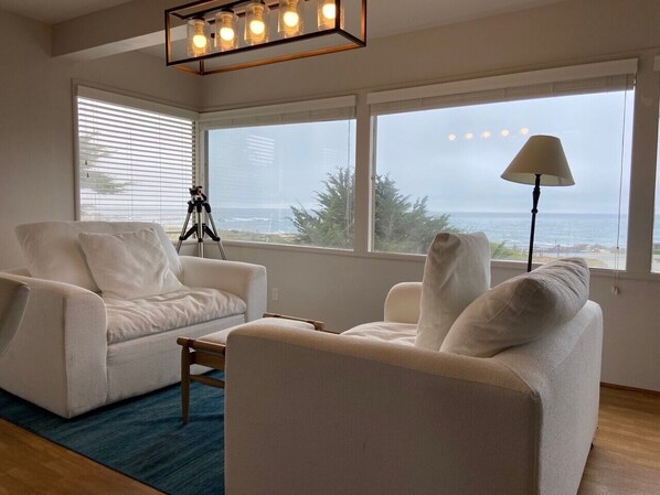 A living room with two white sofas, a coffee table, a tripod, and a lamp. Large windows offer a view of the ocean and buildings. - On the water in Pacific Grove (Pacific Grove)