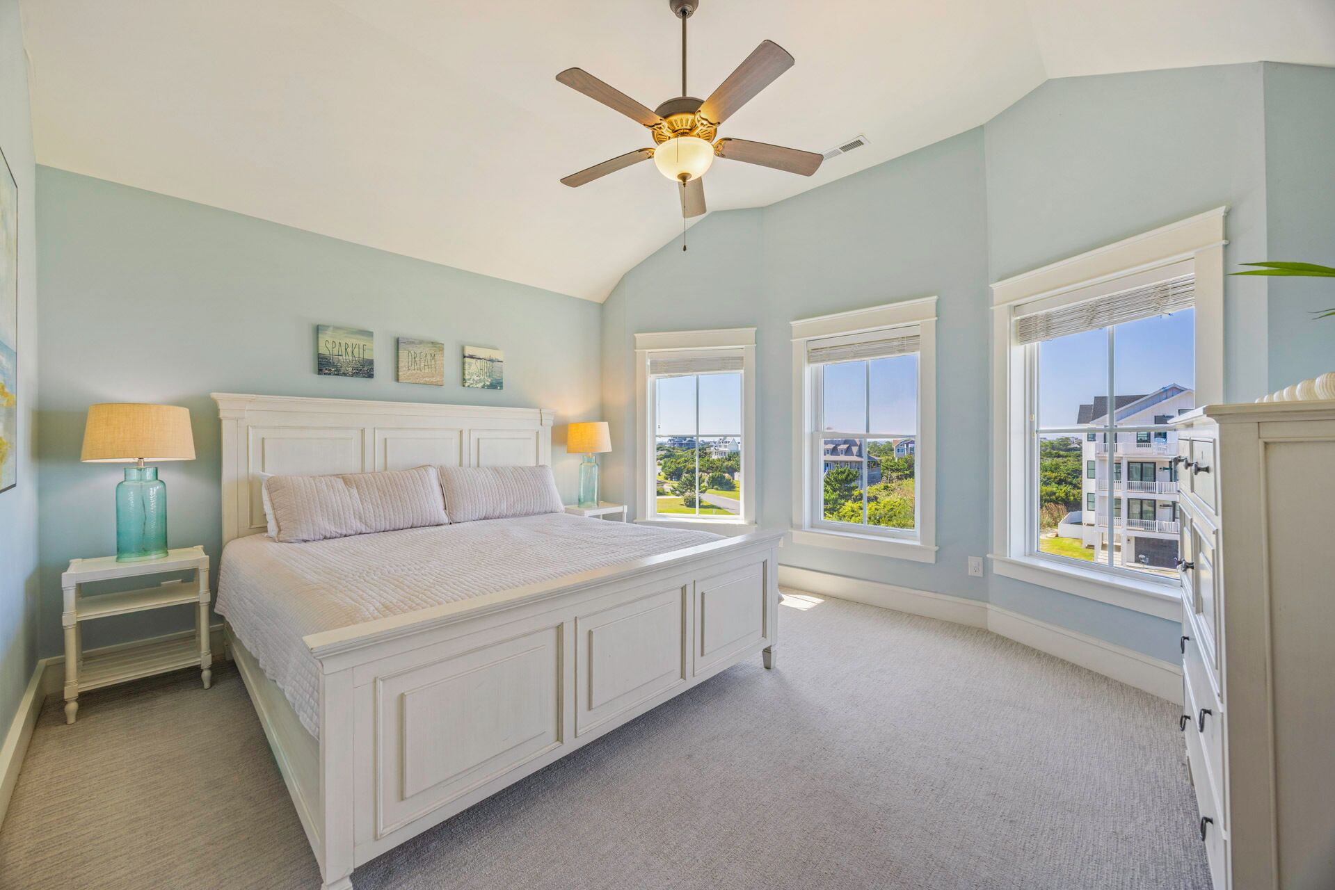 A bedroom with a bed, nightstands, a ceiling fan, and large windows offering a view of the neighborhood.