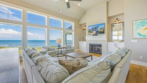 A spacious living room with a large window overlooking the ocean, a fireplace, and a cozy seating area. - Premier Oceanfront w Pool, Hot Tub, Elevator, Theater, Game Room, Outdoor Lounge (Hatteras)