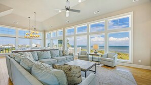 A spacious living room with a large sofa, a coffee table, and a view of the ocean through large windows. - Premier Oceanfront w Pool, Hot Tub, Elevator, Theater, Game Room, Outdoor Lounge (Hatteras)