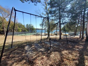 A playground with swings and a tennis court, surrounded by trees and a view of a lake.