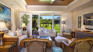 A spacious living room with a large sliding glass door leading to a lush garden, wicker furniture, and a painting of a beach scene. - The Islands at Mauna Lani - Hikialani Residences B2 - Walk to beach and shops (Kamuela)