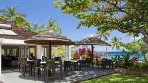 An outdoor dining area with thatched umbrellas, tables, and chairs, overlooking a scenic ocean view. - The Islands at Mauna Lani - Hikialani Residences B2 - Walk to beach and shops (Kamuela)