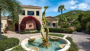 Une fontaine ornée d’une sculpture centrale, un bâtiment doté d’un toit en tuiles et un jardin aménagé avec des palmiers.