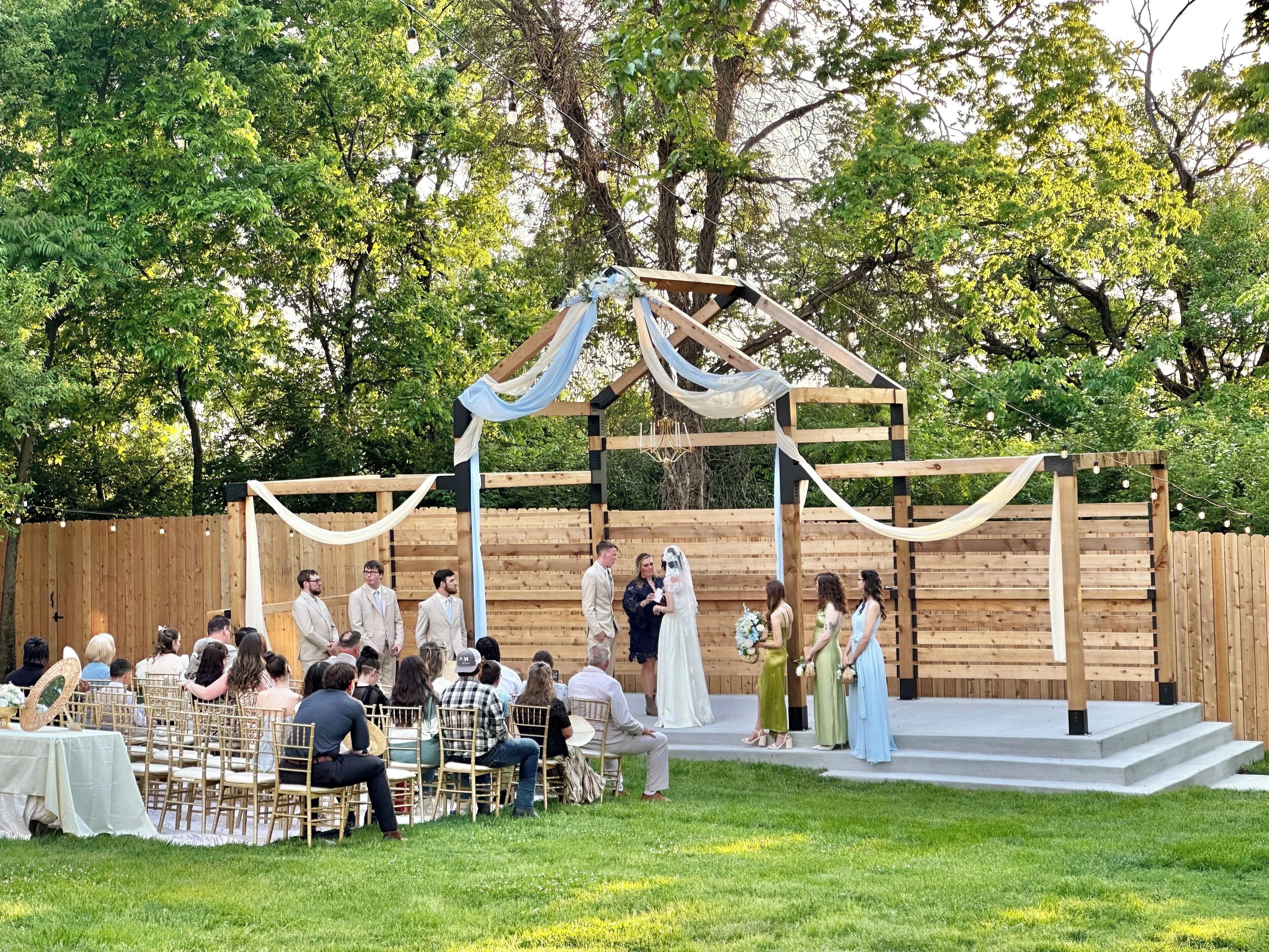 An outdoor wedding ceremony with guests seated on chairs arranged in rows, a wooden stage with draped fabric, and a backdrop of trees.