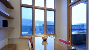 Ein moderner Essbereich mit einem Glastisch, Holzregalen und großen Fenstern mit Blick auf Gebäude und Hügel.