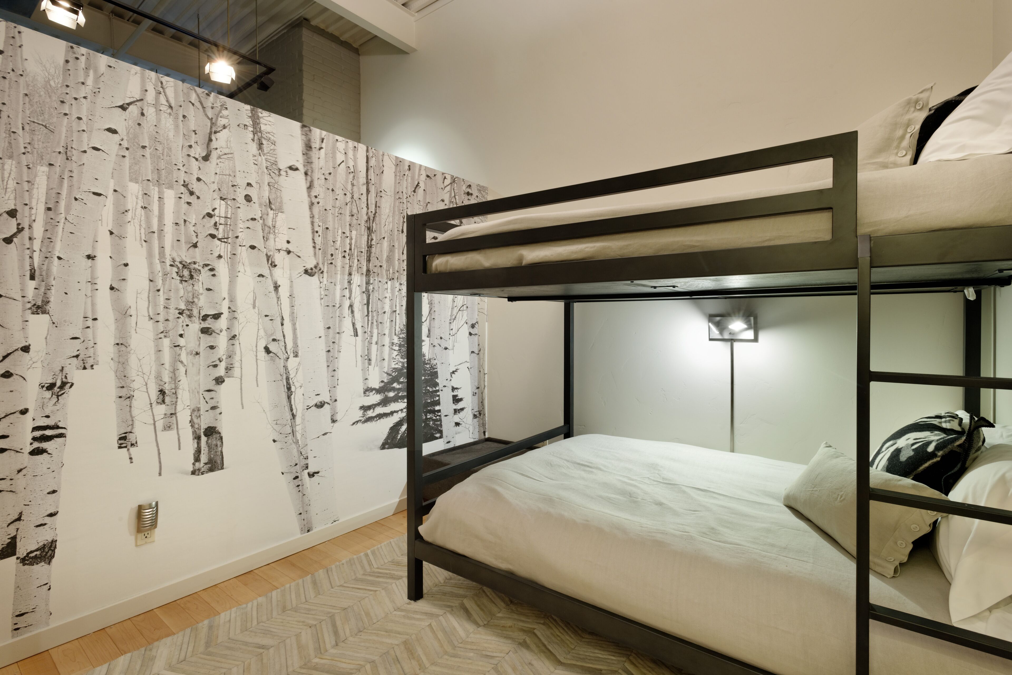 A bunk bed room with a birch tree wallpaper, a wooden herringbone floor, and a wall-mounted light fixture.