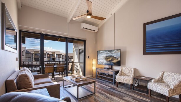 A spacious living room with a large window, a ceiling fan, a flat-screen TV, and a view of the ocean. - Steps to Kamaole 3 Beach (Kihei)