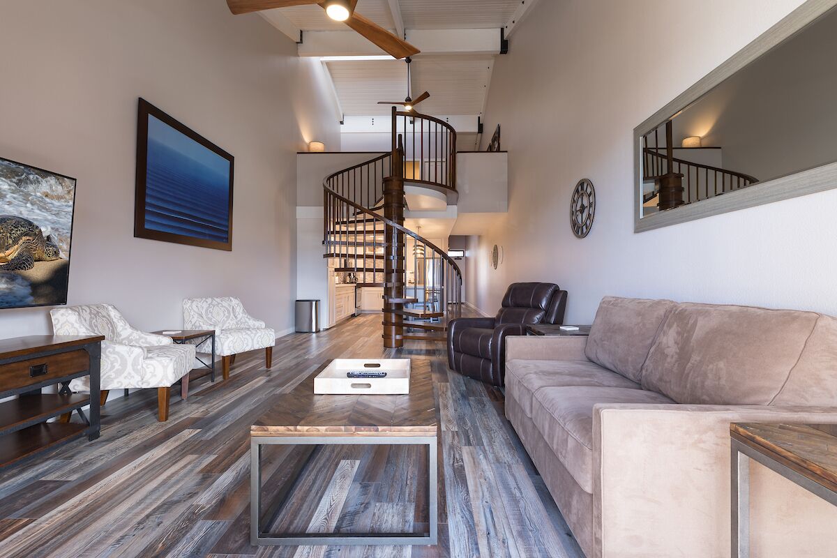 A spacious living room with a wooden coffee table, a beige sofa, and a leather armchair. A spiral staircase leads to an upper level.