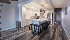 A modern kitchen with a large island, bar stools, and a stainless steel trash can. - Steps to Kamaole 3 Beach (Kihei)