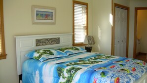Une chambre à coucher avec un lit orné d’un motif représentant une plage tropicale, une table de chevet avec une lampe et un tableau encadré accroché au mur.
