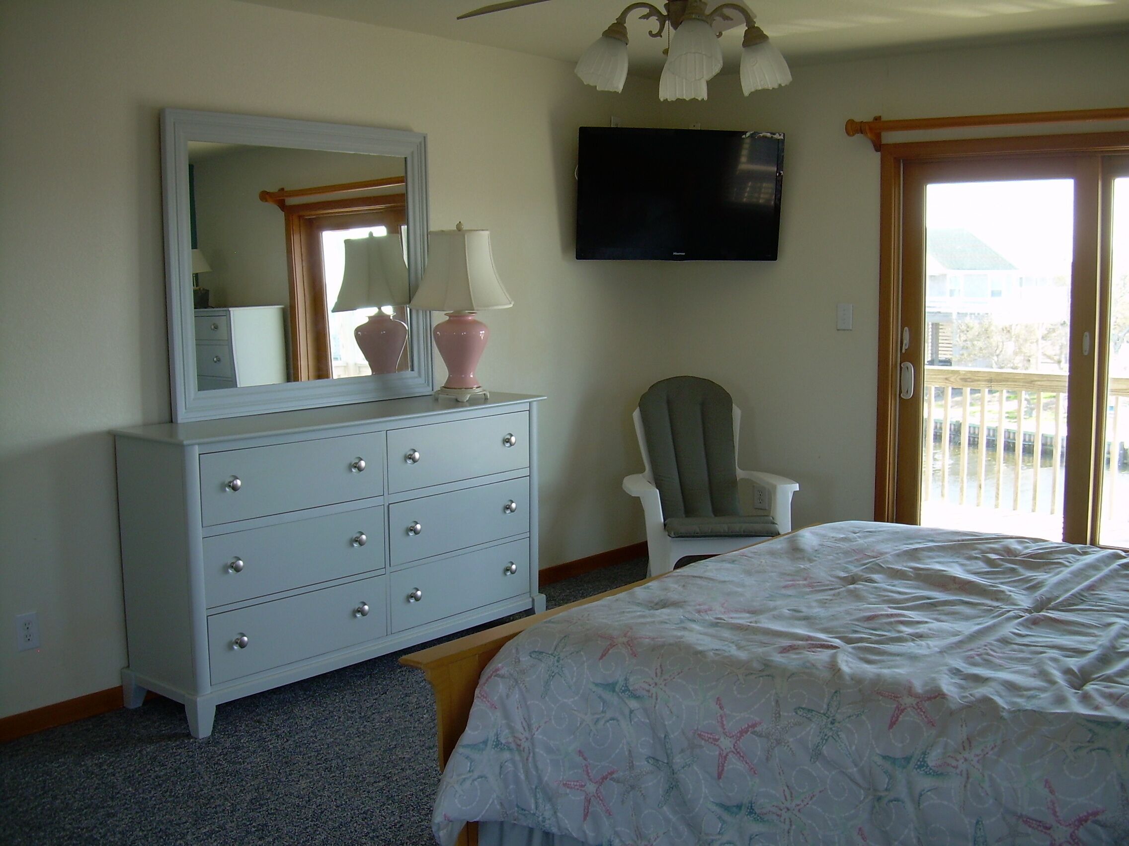 Een slaapkamer met een bed, een dressoir met lades, een stoel, een televisietoestel en een balkondeur.