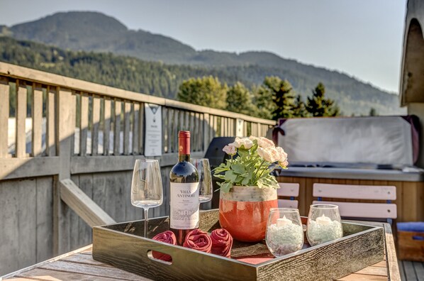 A table with a wine bottle, glasses, a flower arrangement, and candles on a terrace with a mountain view.