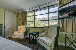 A hotel room with a bed, two armchairs, a small table, and a large window offering a mountain view.