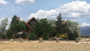 A rustic cabin with a prominent chimney, surrounded by a dry grassy field and various trees.