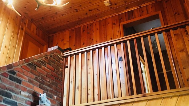 A wooden cabin interior with a brick fireplace, a wooden balcony, and a deer antler chandelier.