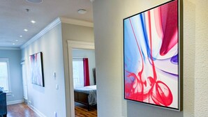 Um corredor com piso de madeira, uma cama ao fundo e duas pinturas abstratas nas paredes.