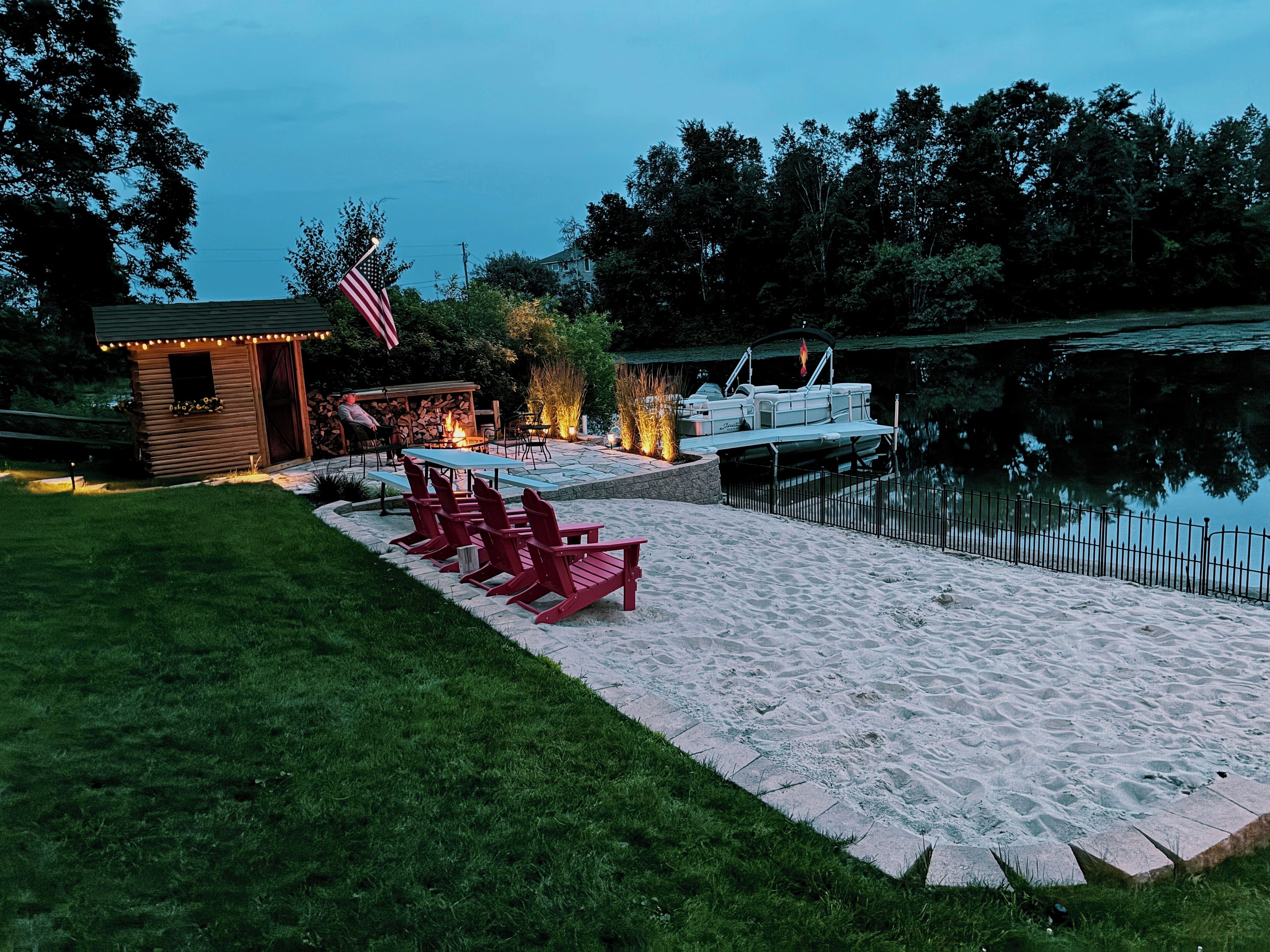 Une terrasse au bord du lac, avec des chaises Adirondack rouges, un petit chalet en bois et un quai avec un bateau.