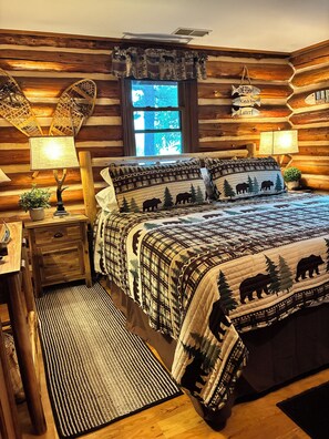 A log cabin bedroom with a bed, two bedside tables with lamps, a window, and a view of the outdoors. - Authentic Log Cabin on Beautiful Silver Lake in Traverse City MI. Pet Friendly! (Traverse City)