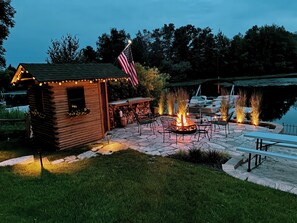 A wooden cabin with a fire pit and outdoor seating, surrounded by a stone patio and a lake. - Authentic Log Cabin on Beautiful Silver Lake in Traverse City MI. Pet Friendly! (Traverse City)